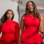 Two women in matching red outfits pose confidently indoors for a fashion or event photo.