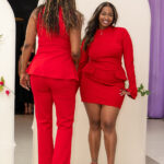 Two women in red outfits stand by a white arched backdrop; one faces the camera smiling, the other has her back to the viewer.