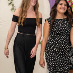 Two smiling young women walk side by side at an event, one in a black off-shoulder top and pants and the other in a black polka-dot dress, with a purple backdrop and floral arches behind them.
