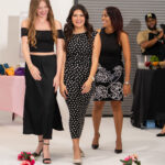 Three women in black outfits walk a white runway, smiling, with pink flowers lined along the edge.