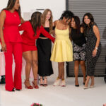 Six women in colorful outfits stand together, smiling for a photo at a fashion event with pink flowers on the floor ahead of them.