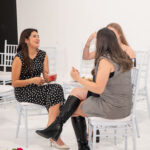 Three women seated on white chairs in a bright room, smiling and chatting; one holds a pink cocktail.